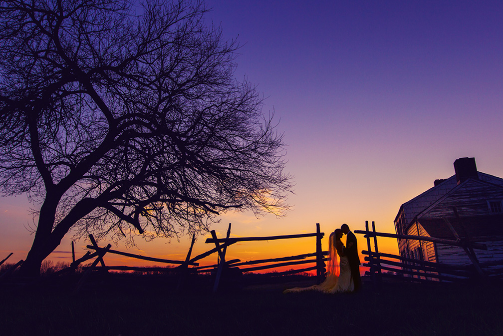 Silhouetted couple embracing by a wooden fence and old barn under a purple and orange sunset