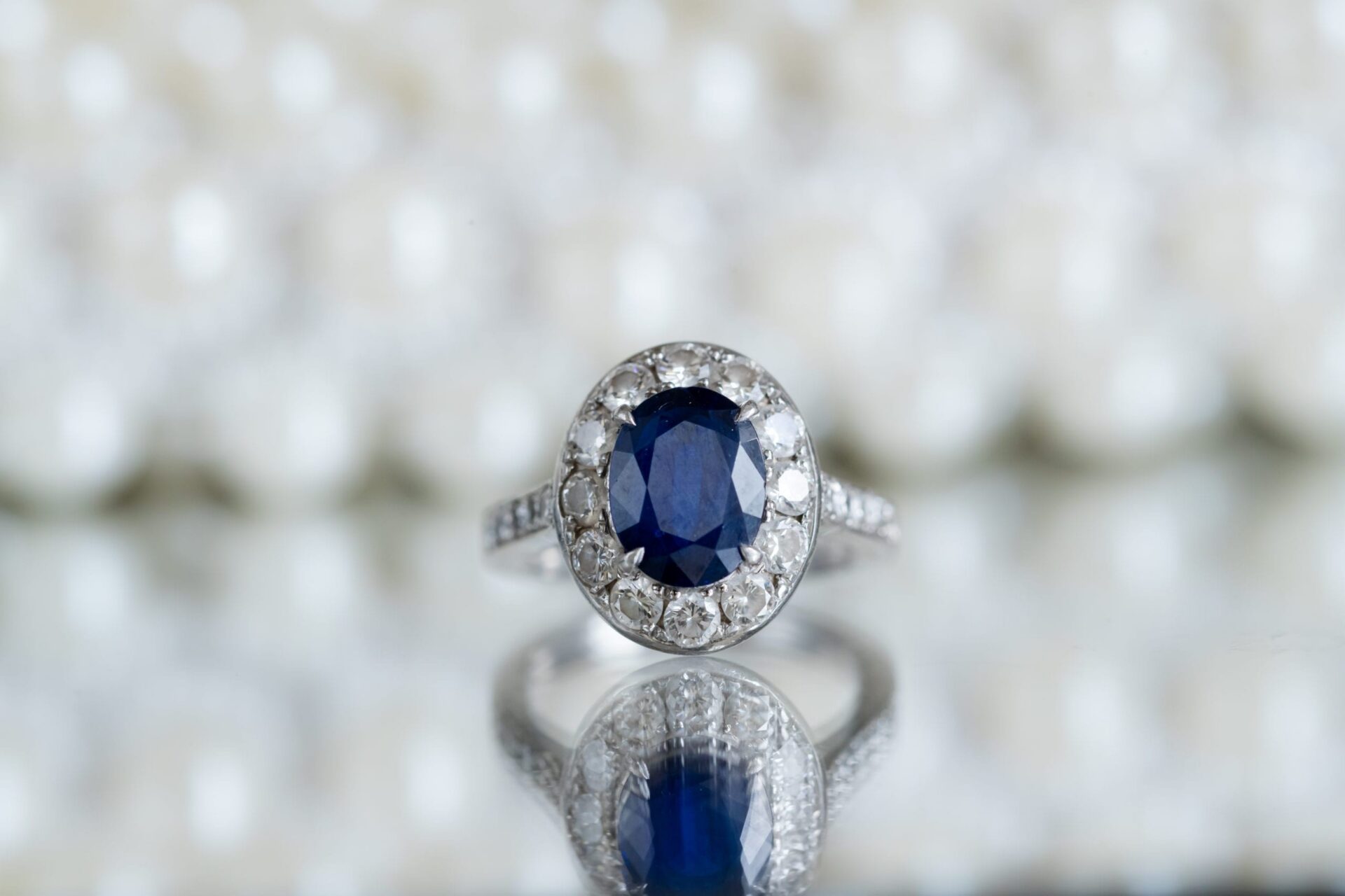 Oval blue sapphire halo ring on a mirrored surface with blurred pearls