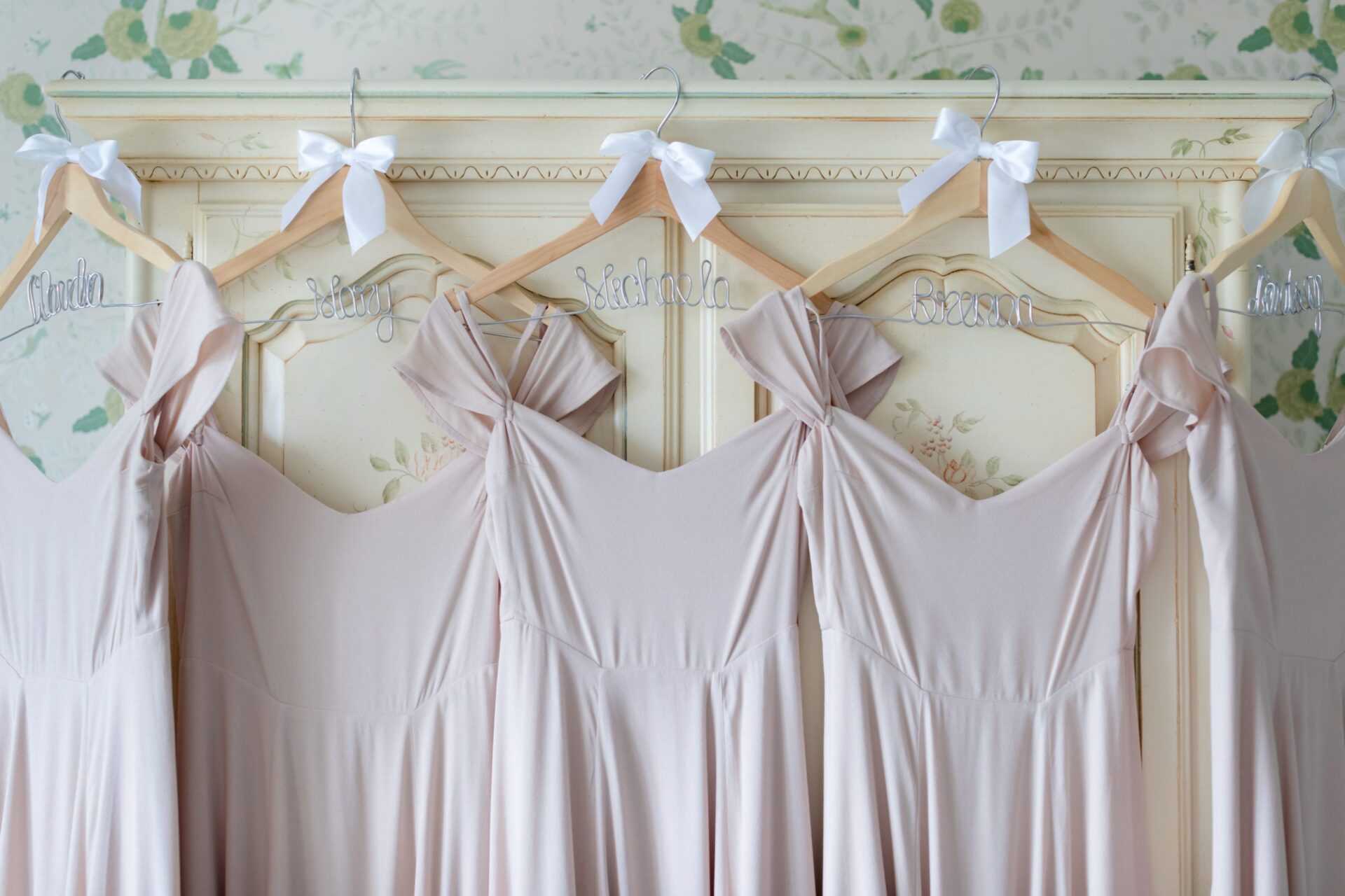 Blush bridesmaid dresses on name hangers with white bows against a painted cabinet