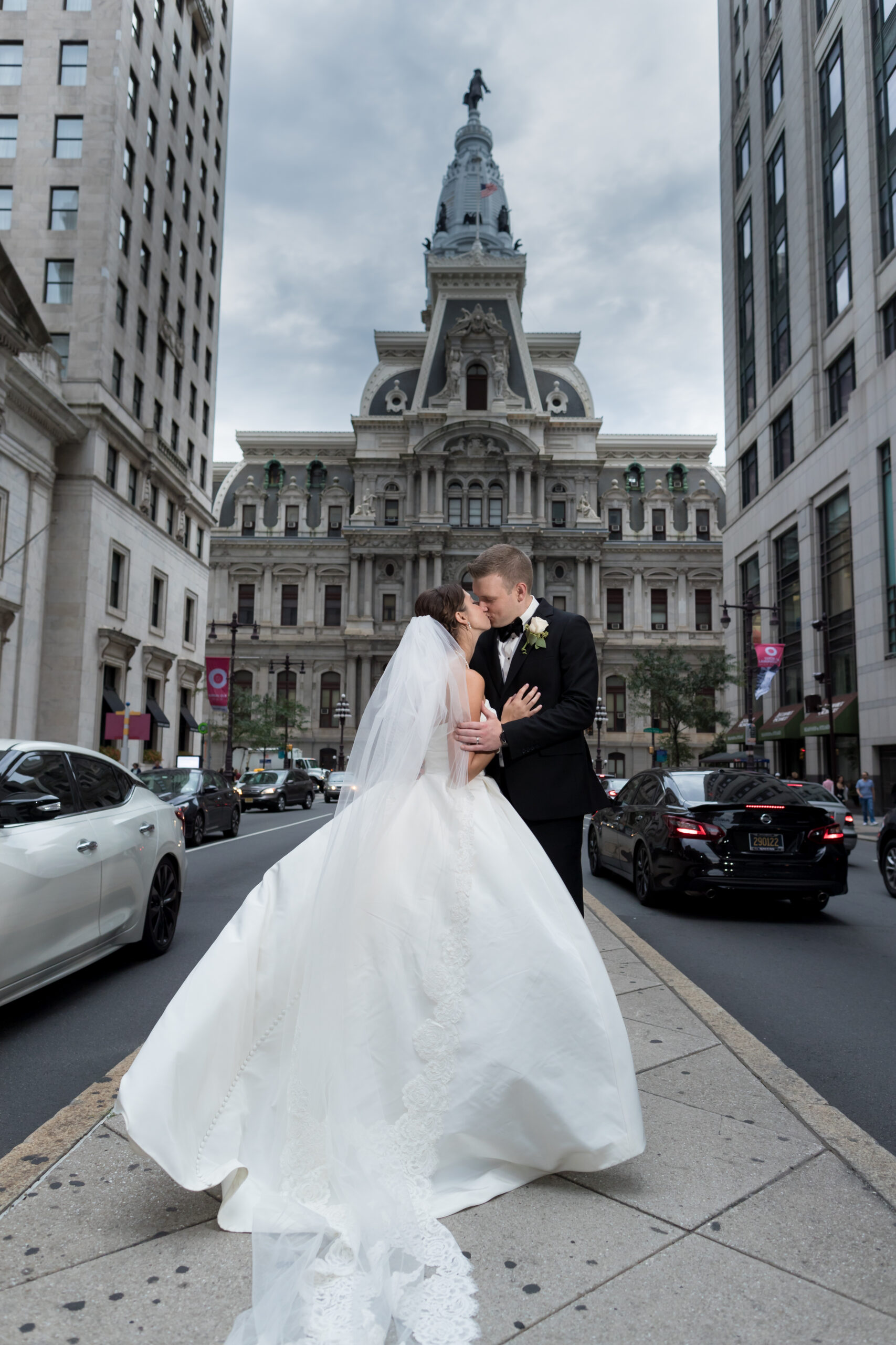 Bride and groom kissing on a city street with her long train flowing in front of Philadelphia City Hall