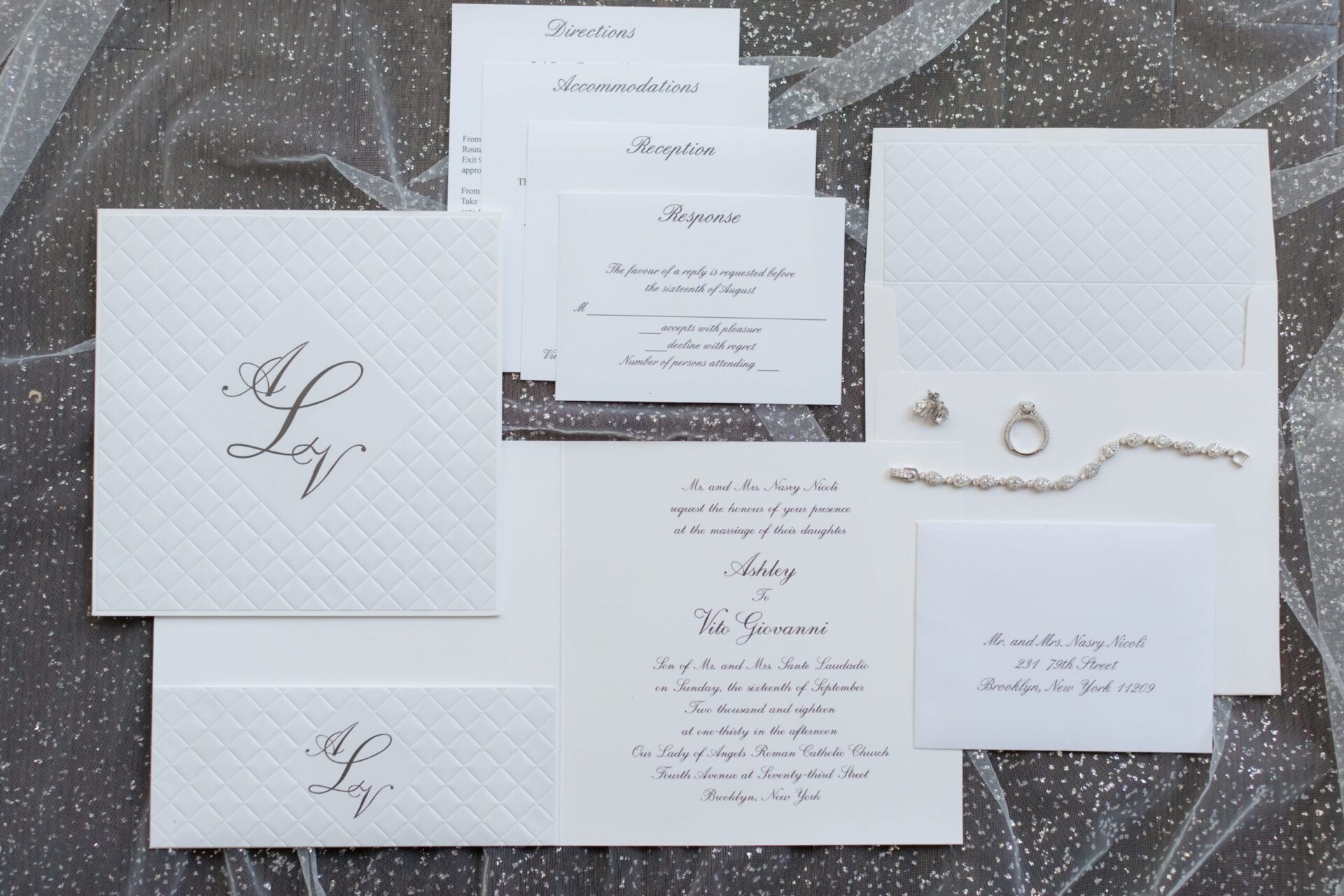 Embossed white invitation suite with a script monogram, ring and pearl bracelet on glitter tulle