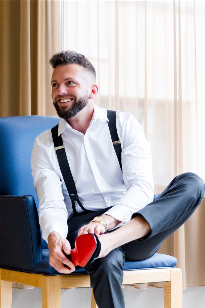 Groom in suspenders sitting on a blue chair putting on a shoe with a red sole