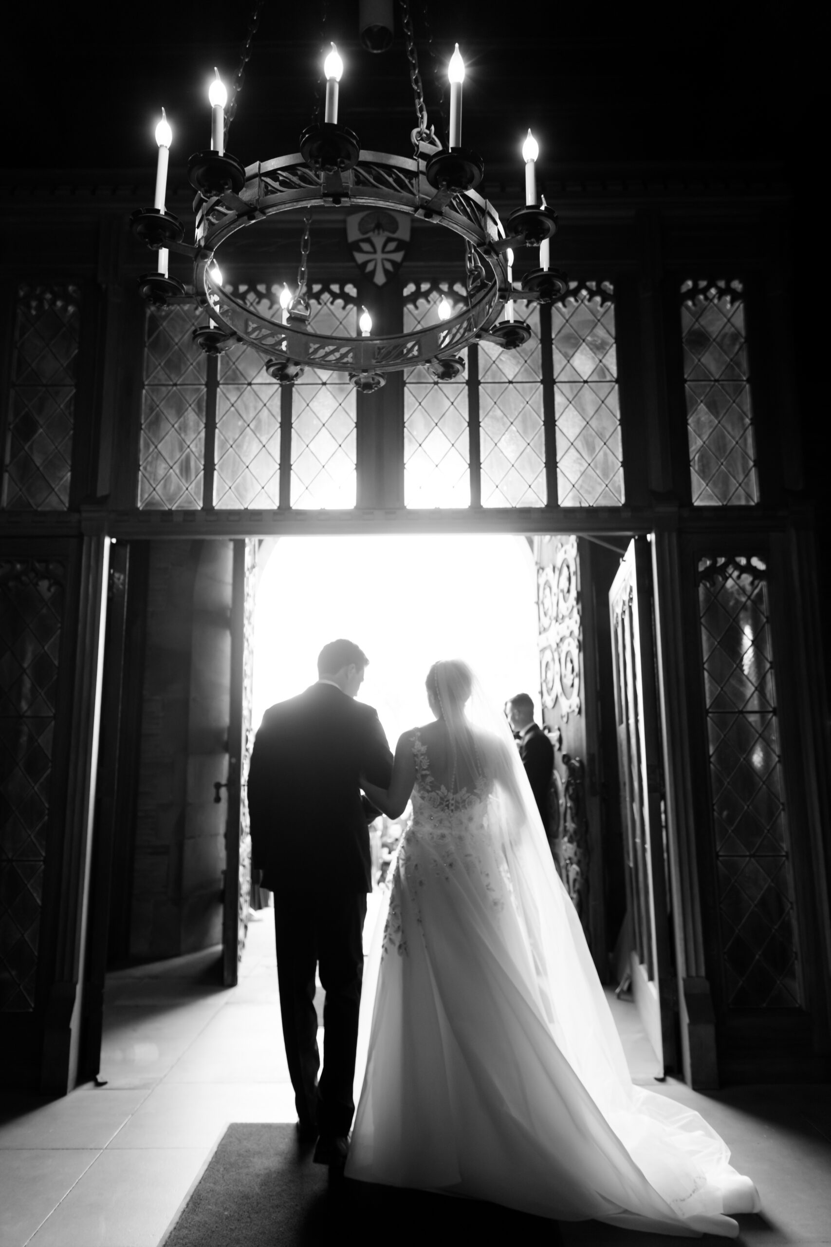 Black and white photo of the bride and groom walking out through open chapel doors under a chandelier