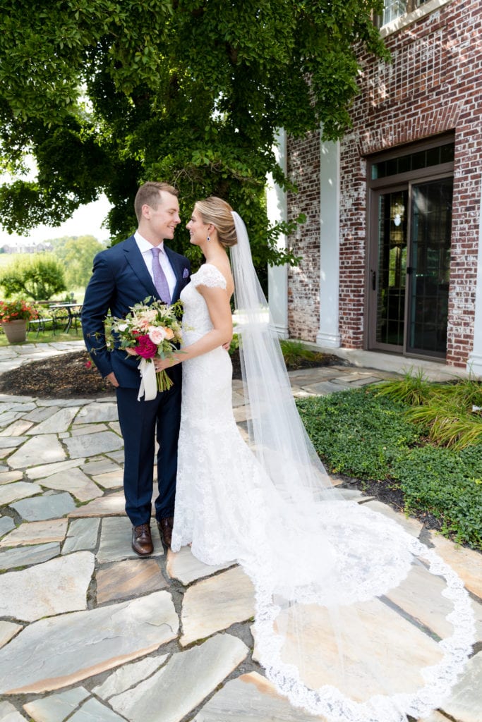 Bride in a lace gown with a long veil and groom in navy on a flagstone patio beside a brick building