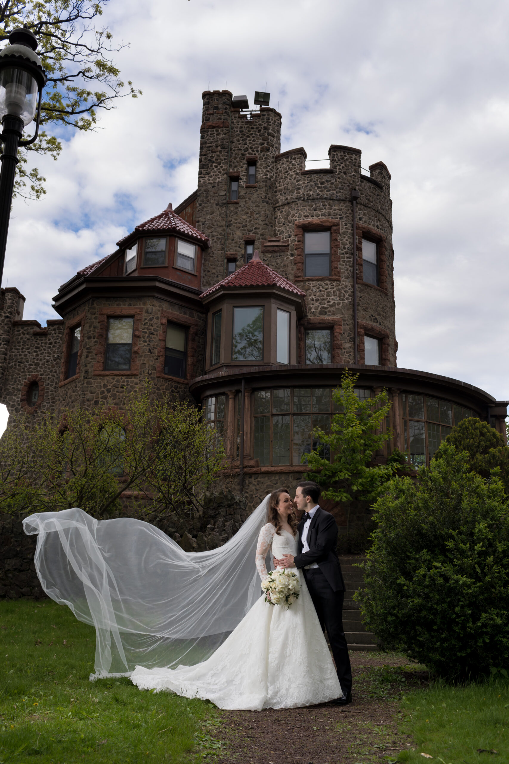 Bride and groom kissing on the lawn with her long veil blowing in front of a stone castle