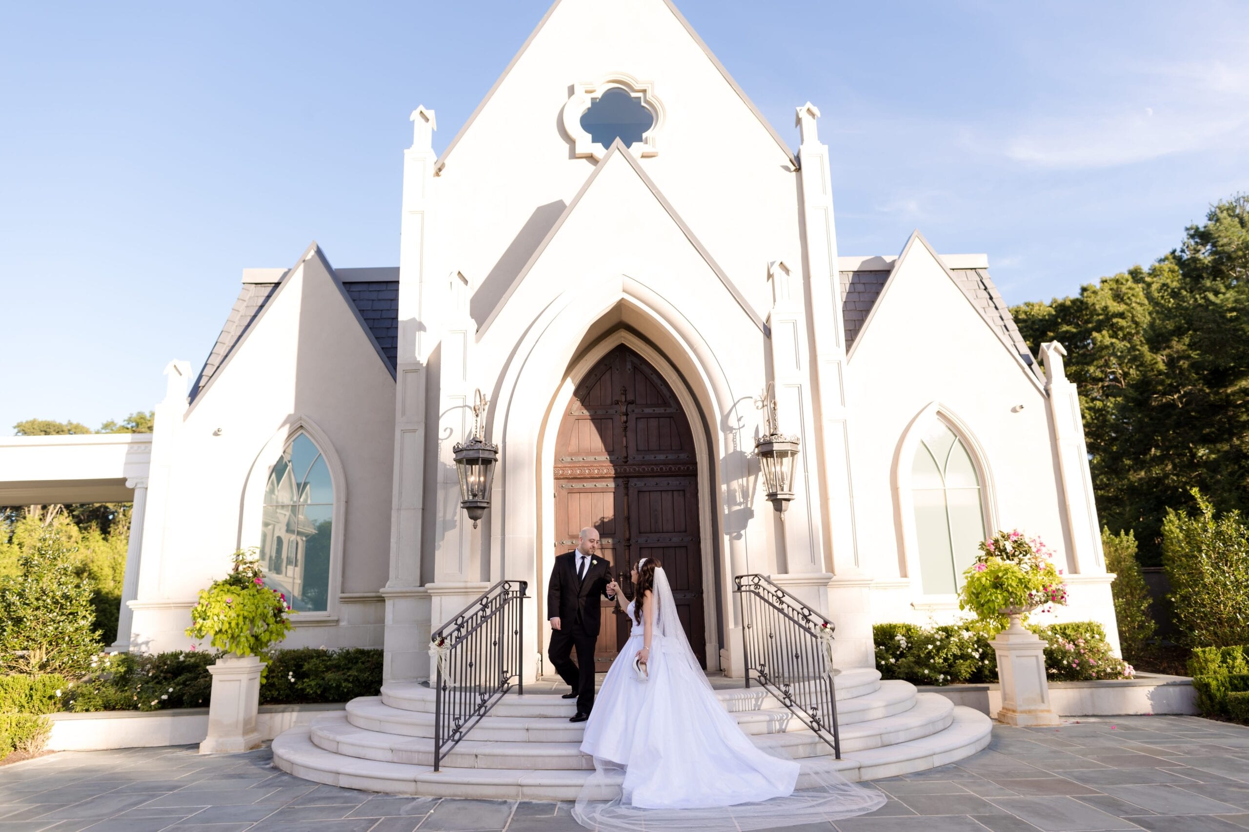 Bride in a ball gown and a man in a tuxedo walking down the steps of a white stone chapel