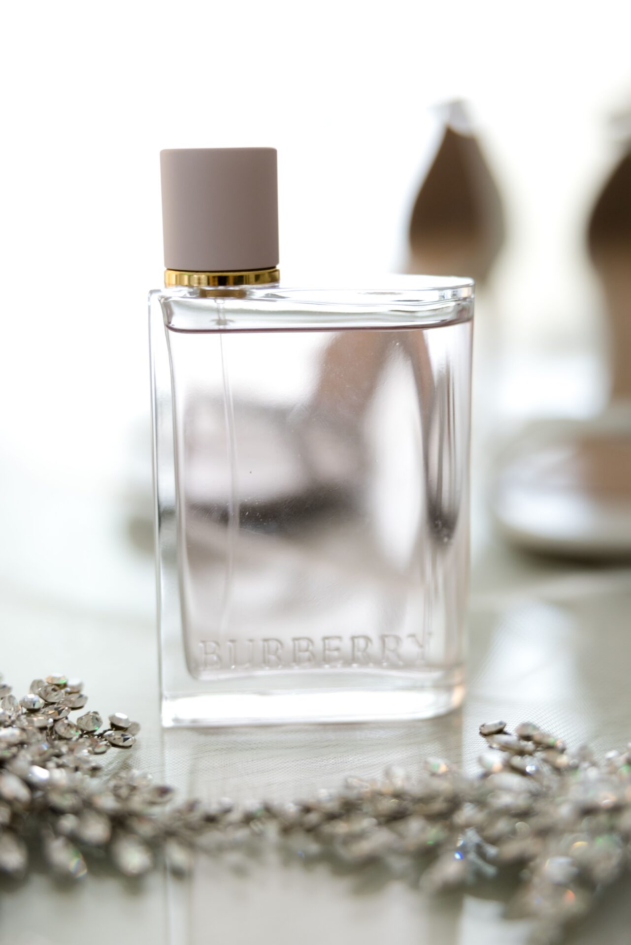 Clear perfume bottle among scattered crystals with blurred white heels behind