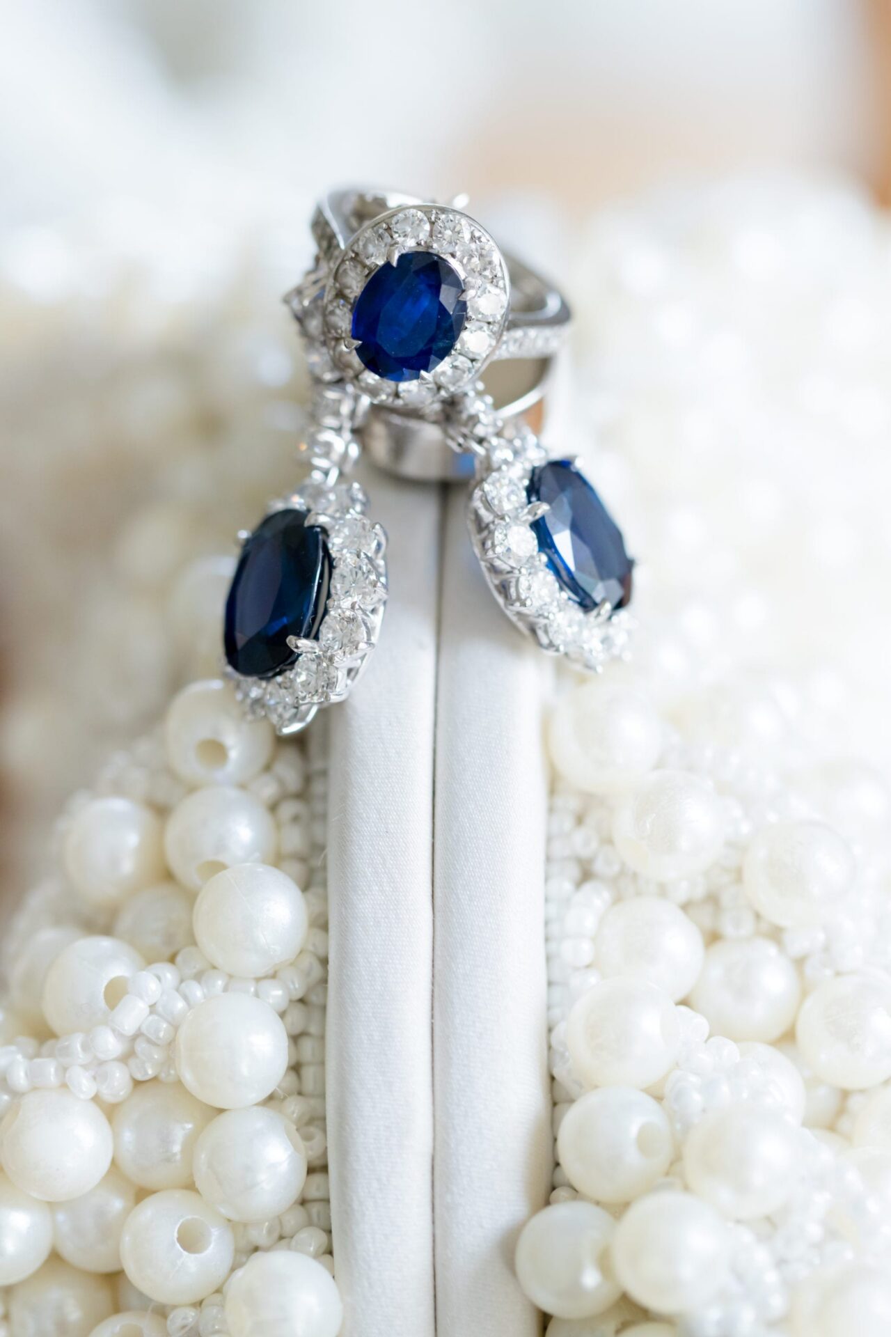 Sapphire halo ring and drop earrings displayed on a pearl-beaded clutch