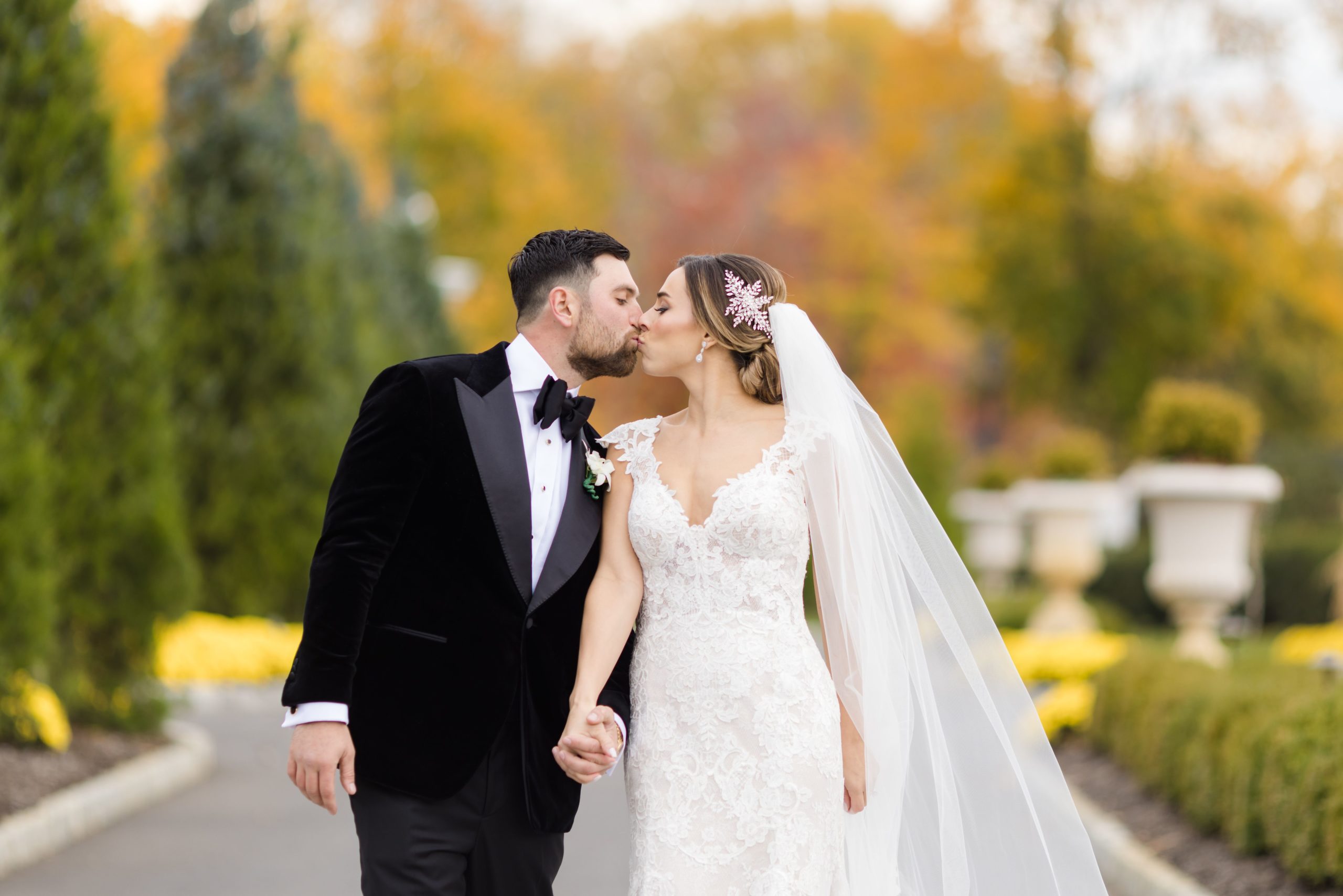 Bride and groom in a velvet tuxedo jacket kissing while holding hands on a path lined with autumn foliage