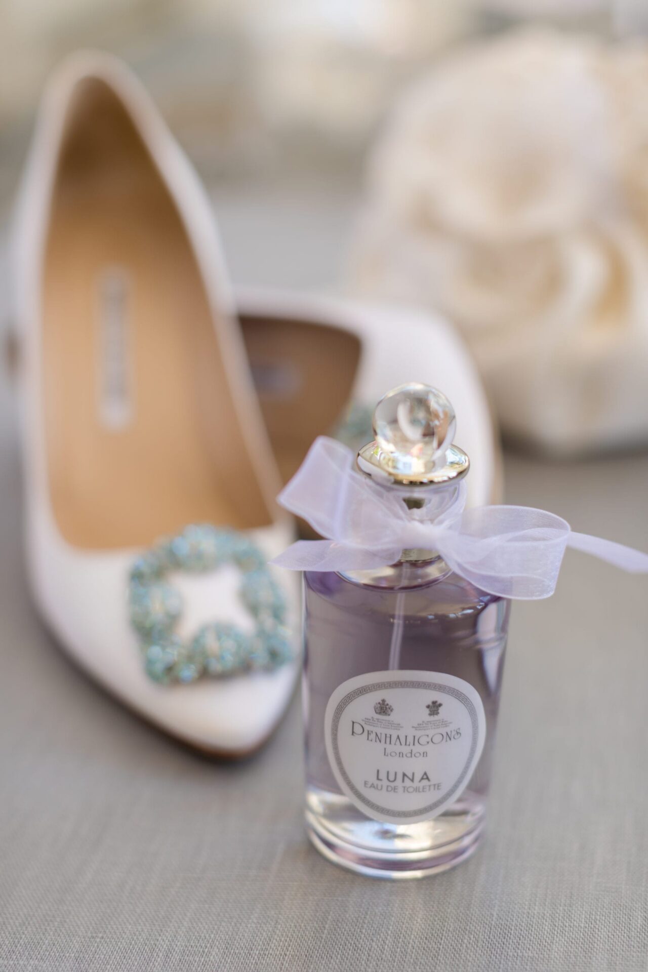 White heels with a turquoise jeweled buckle beside a perfume bottle tied with sheer ribbon