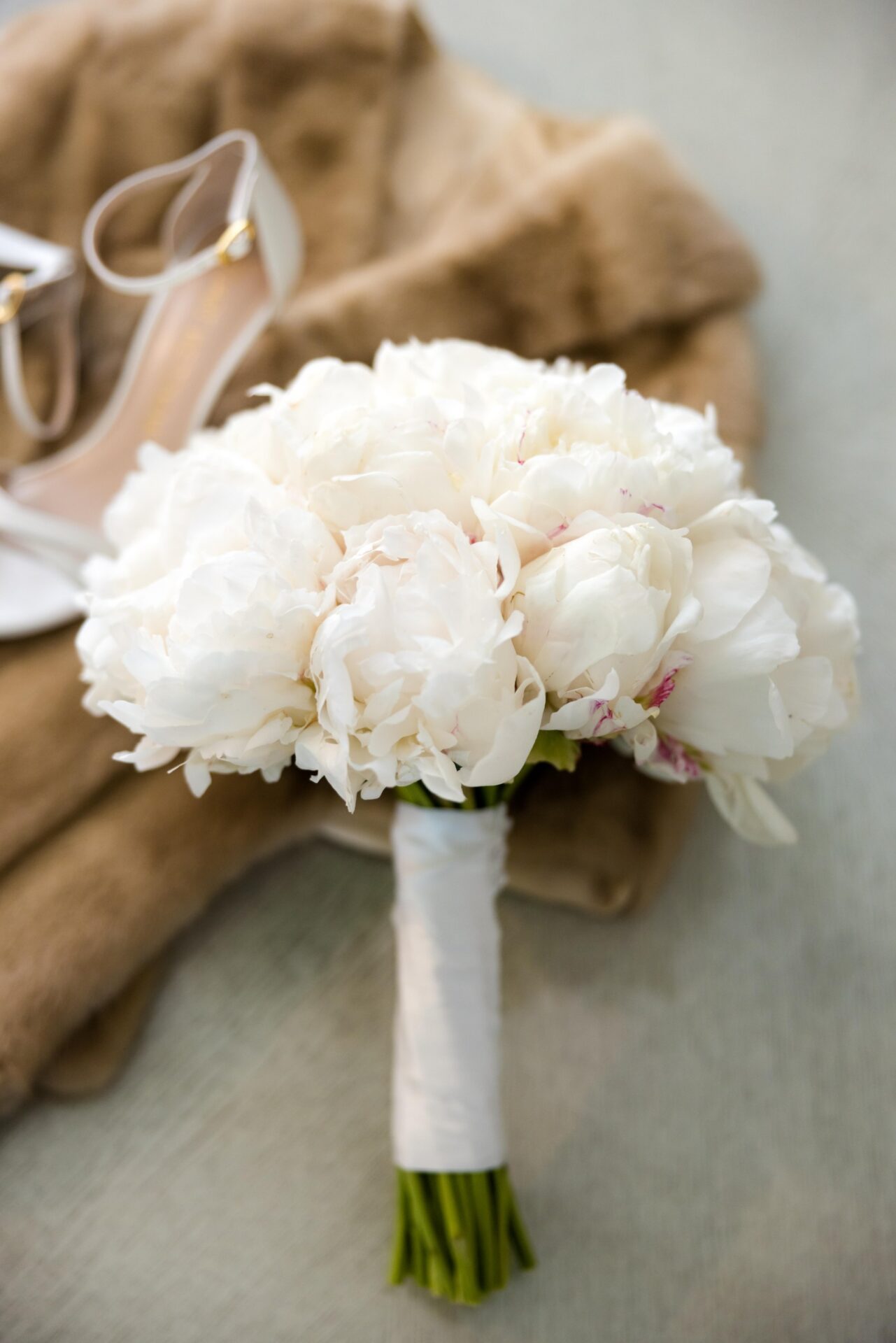 White peony bouquet on a fur wrap beside a white ankle-strap heel